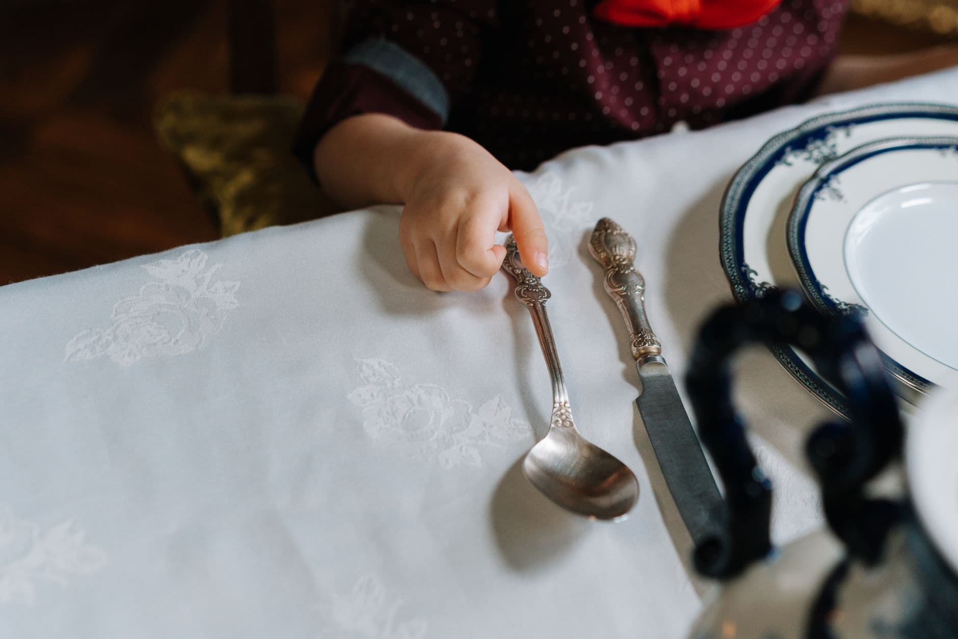 A small child's hand beside a silver spoon and knife on a white tablecloth at a family celebration table