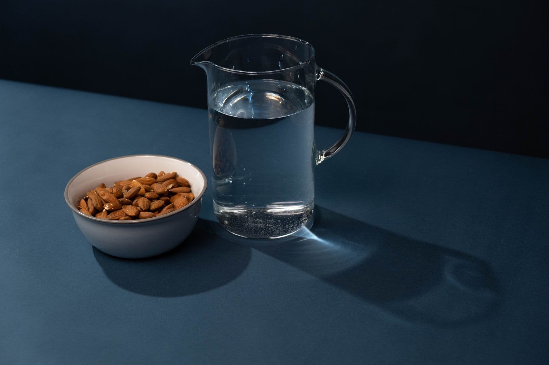 A clear glass pitcher of water beside a bowl of almonds on a calm dark background