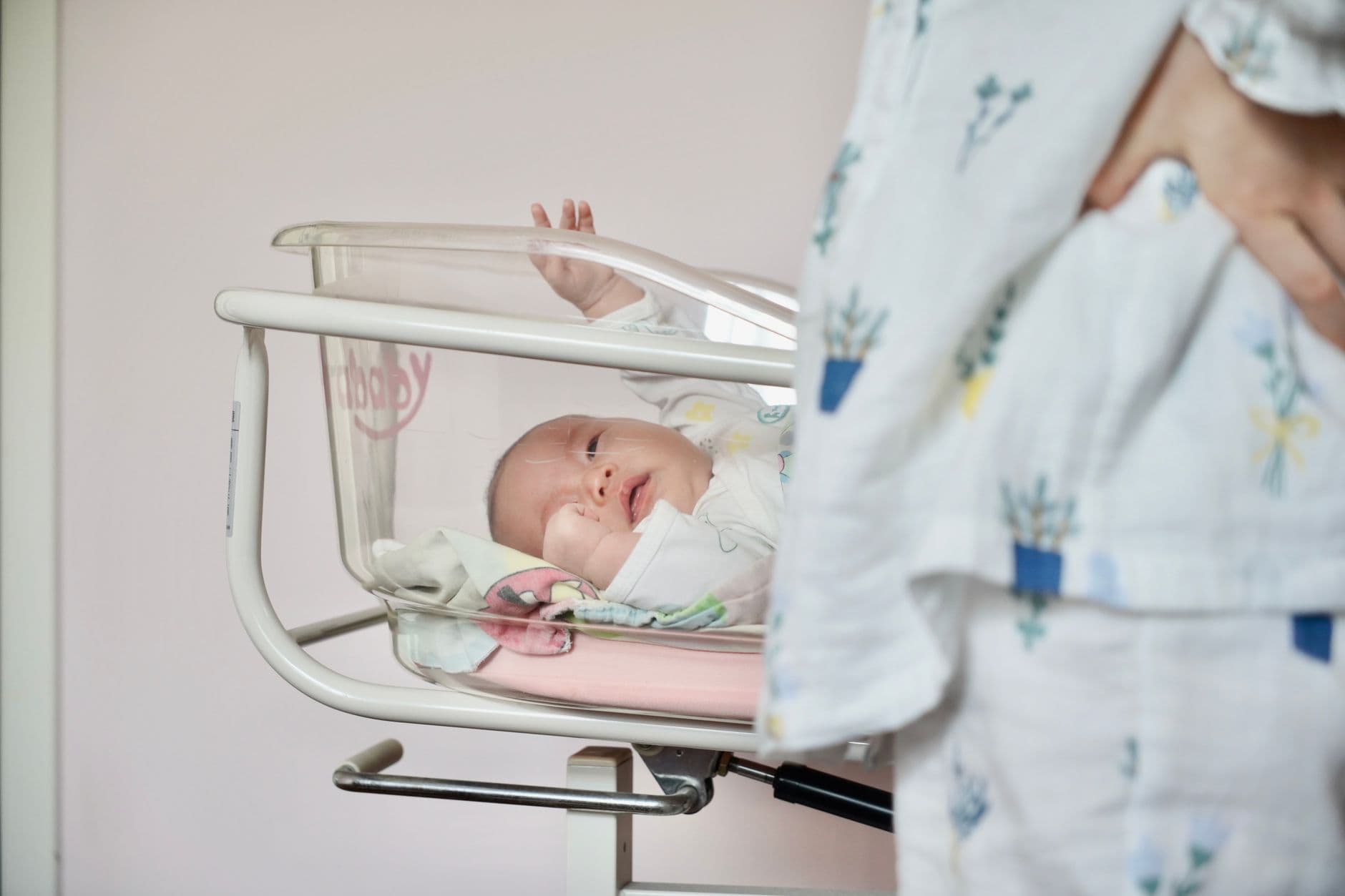 A newborn in a clear hospital bassinet raising one hand, with the mother standing beside it in a hospital gown